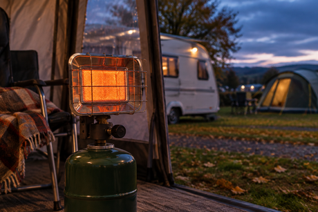 A portable gas heater with a glowing ceramic burner on a green gas cylinder, lit inside an awning at dusk with a caravan and tent in the background